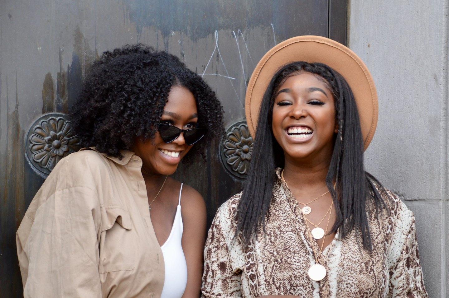 Two smiling Black women stand together outdoors near a grey building.