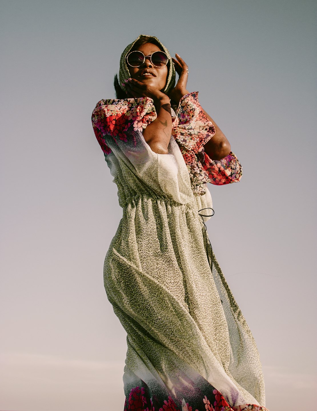 Woman in a patterned floral dress posing outdoors under a clear sky.